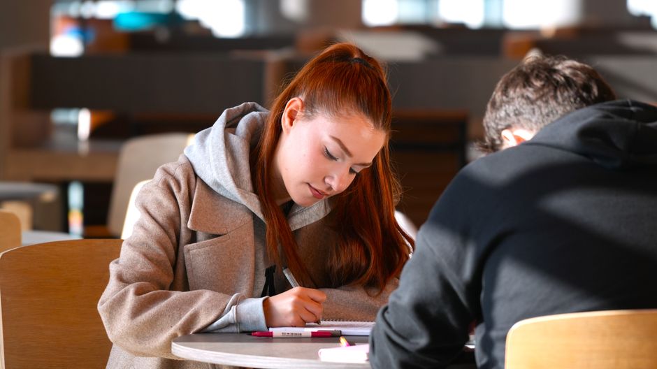 A young woman and man study together at a library table, focused on their notes.