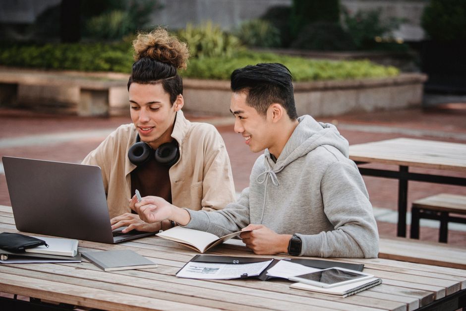 Cheerful diverse young male students in casual outfits sitting at wooden table with gadgets and papers while working together on hometask