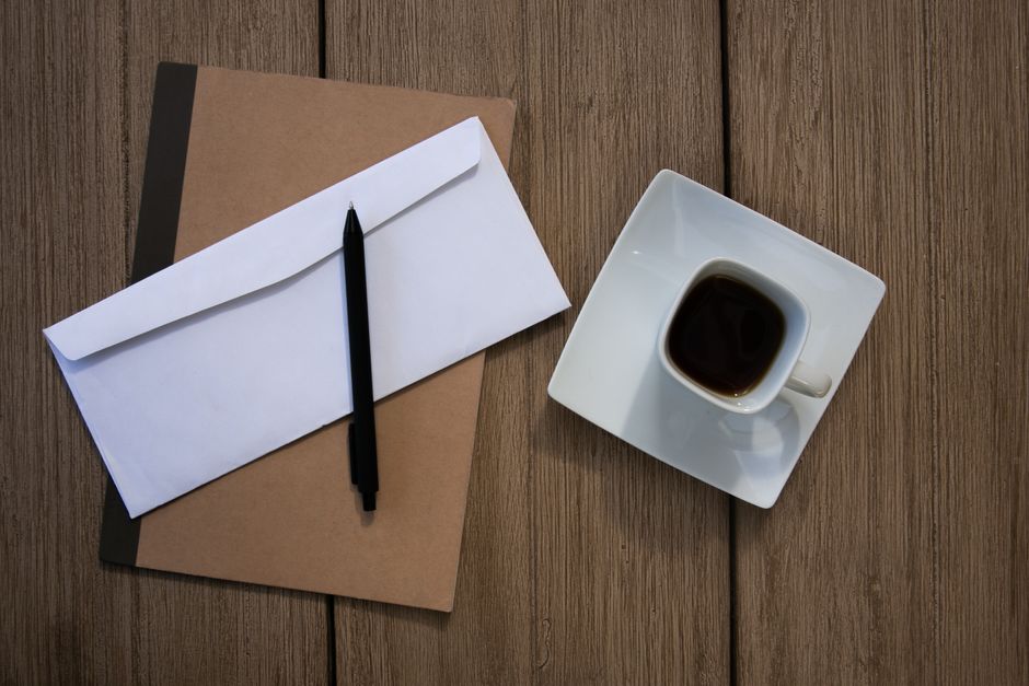A cup of coffee, notebook, envelope, and pen on a rustic wooden table.