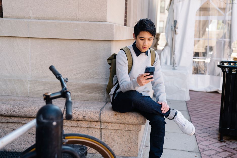 A young student in casual attire uses a smartphone while sitting outdoors on a university campus.