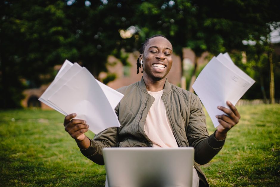 Super positive black student sitting in green park with application papers and laptop and smiling happily after learning news about being accepted to university